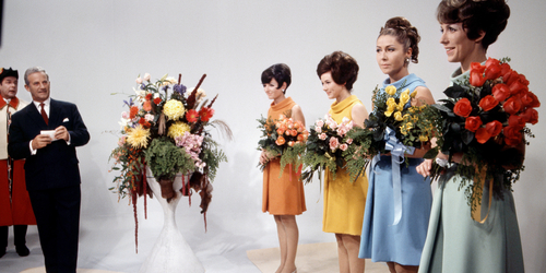 Vier Frauen mit Blumensträussen, eine in einem orangenen Kleid, eine in Gelb, eine in Hellblau und eine in Zartgrün.