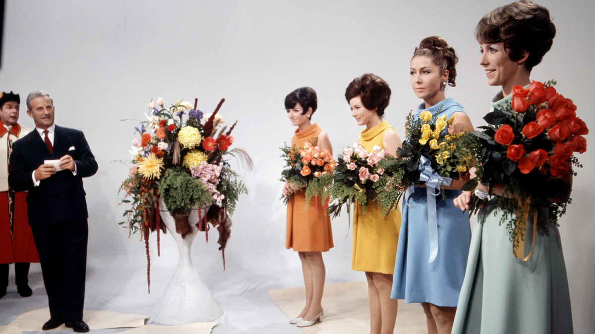 Quattro donne con mazzi di fiori: una con un vestito arancione, una con uno giallo, una con uno azzurro e una con uno verde chiaro.
